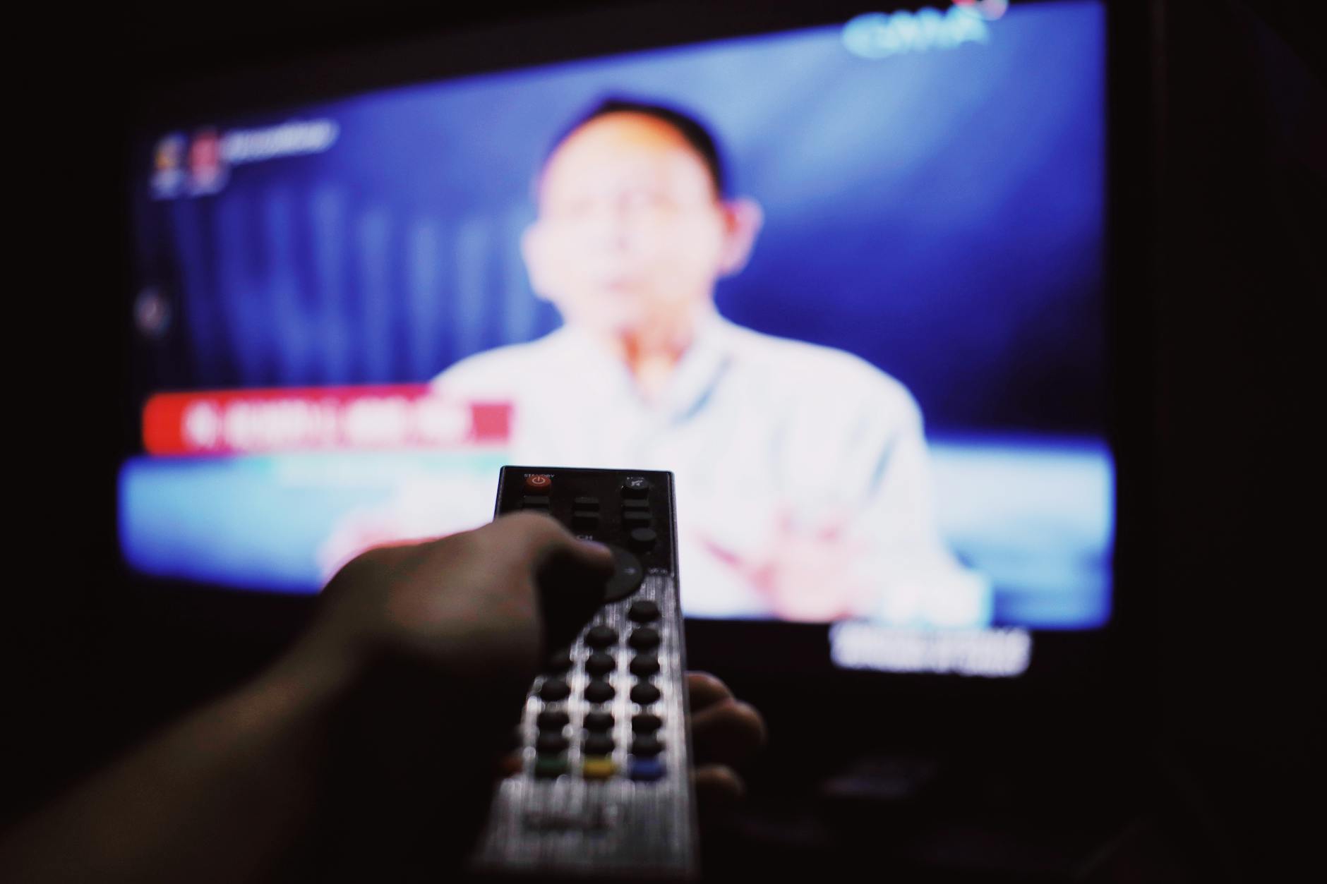 Anonymous viewer holding a remote control in a dim living room, switching channels.