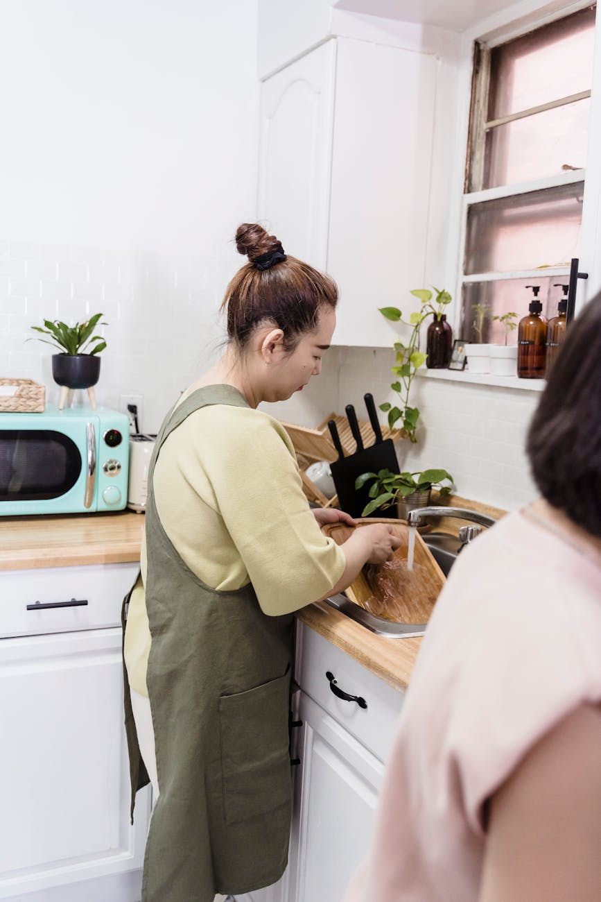 Woman in apron washing a chopping board at a bright kitchen sink