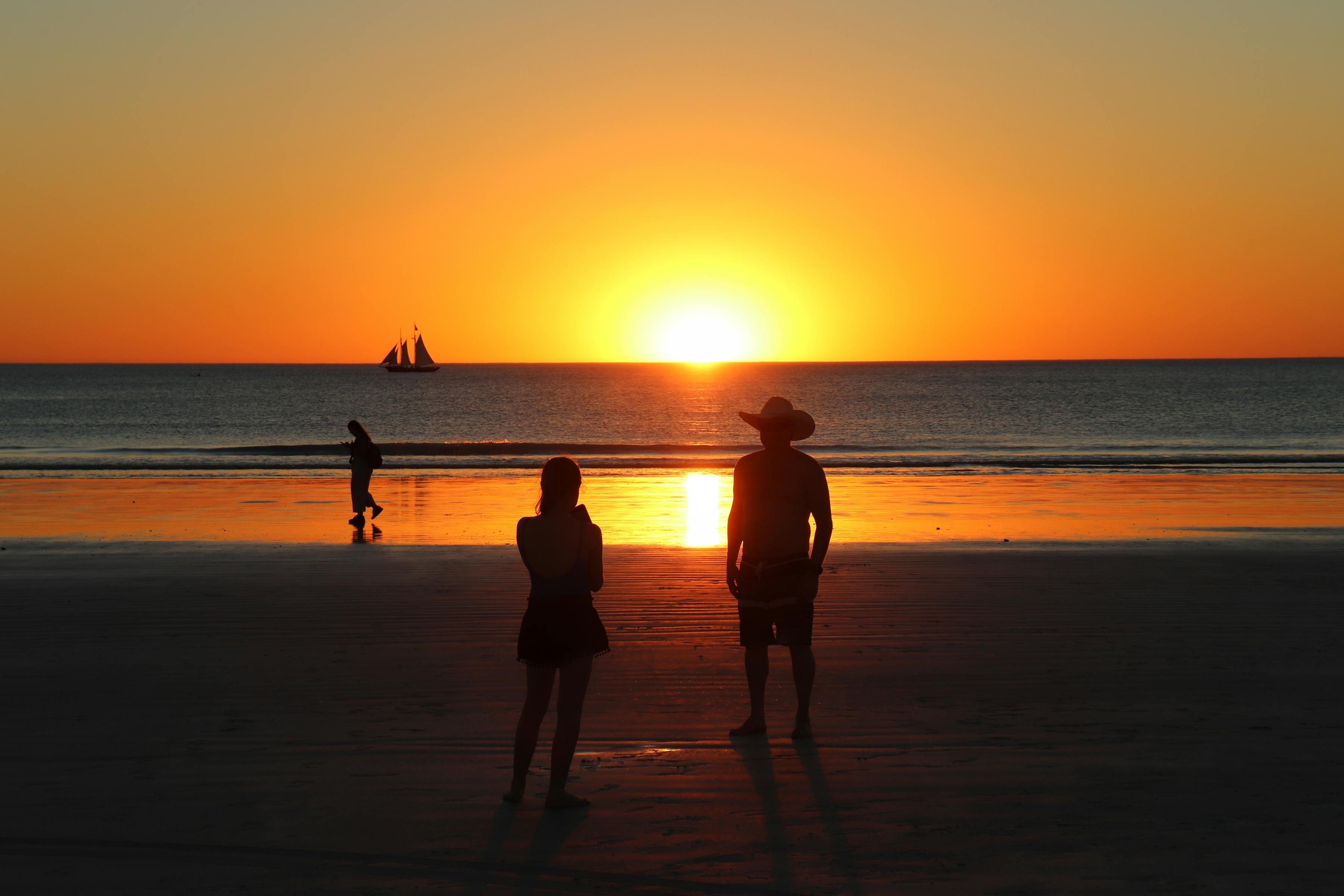 Silhouettes of camels and riders against a vivid orange sunset and sailboat at Cable Beach, Broome, Western Australia