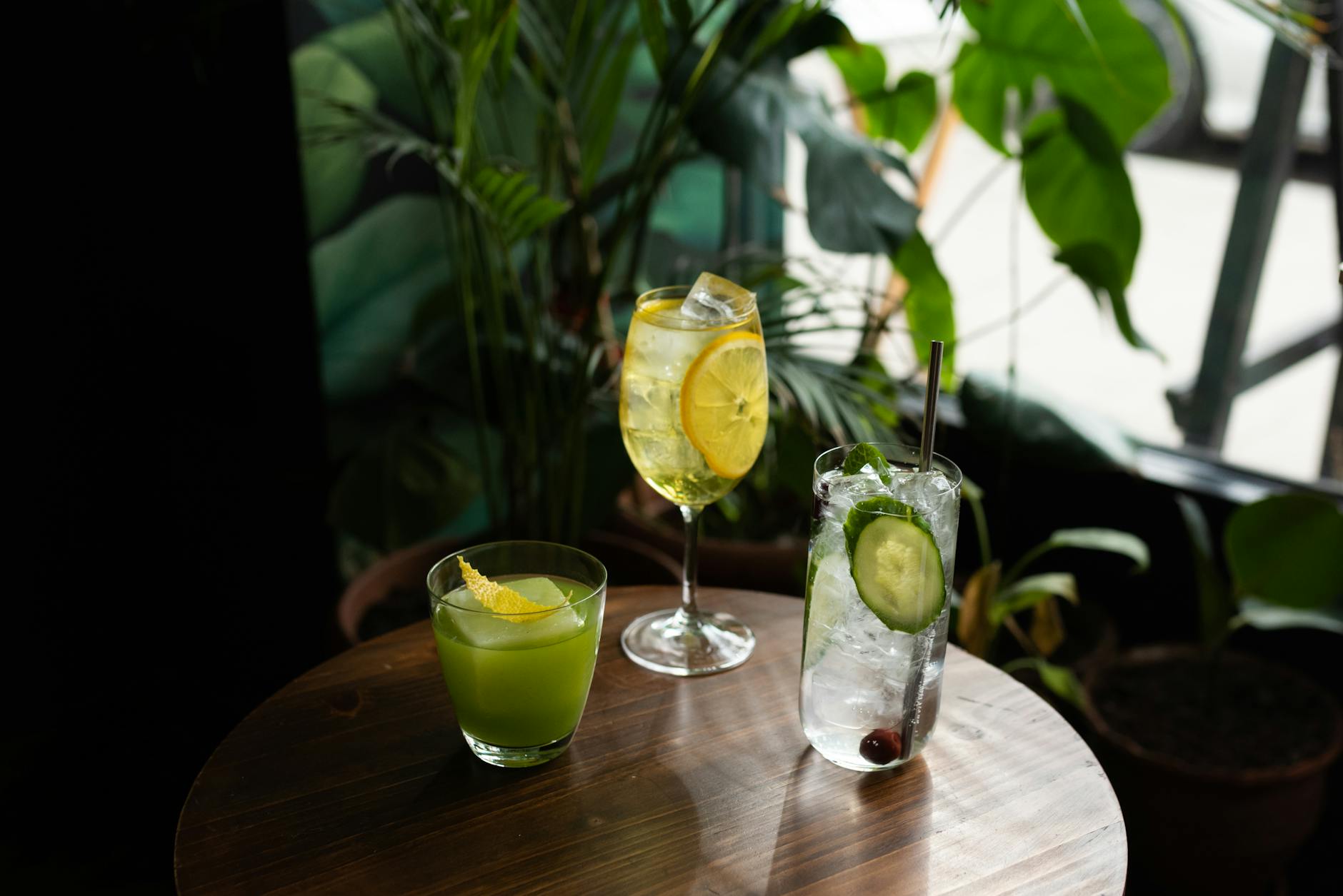 A trio of cocktails featuring lemon and cucumber garnishes on a wooden table surrounded by plants.