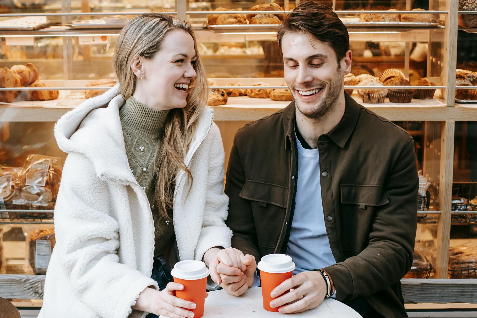 Delighted young couple wearing casual clothes laughing while sitting close together and holding hands near a bakery with coffee in paper cups