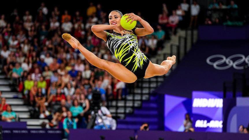 A gymnast does the splits while jumping and holding a ball.