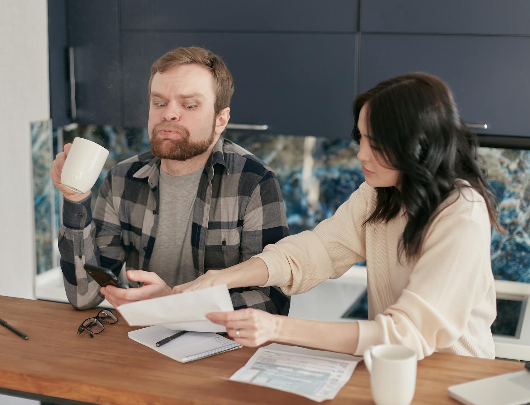 A couple sit at a kitchen table with bills and coffee between them, the ordinary domestic scene where money arguments start.
