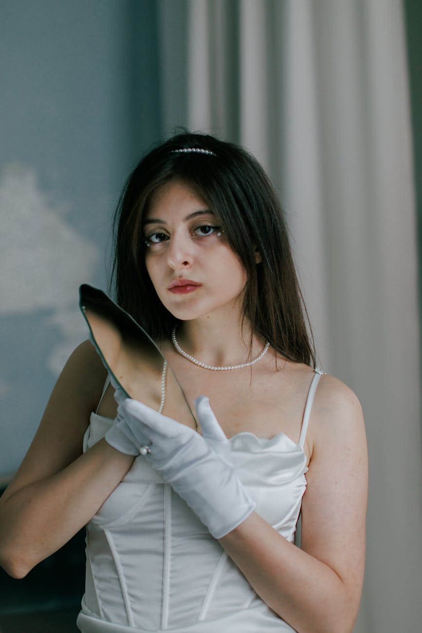 A woman holds a small hand mirror indoors, echoing the self-scrutiny behind the GLP-1 look.