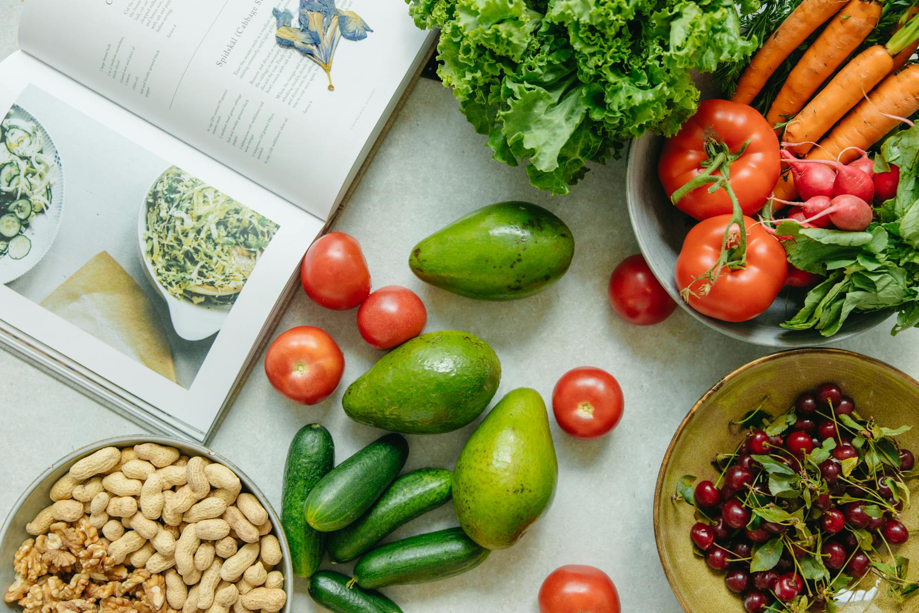 Cookbook beside leafy greens, fruit and whole foods on a kitchen table.