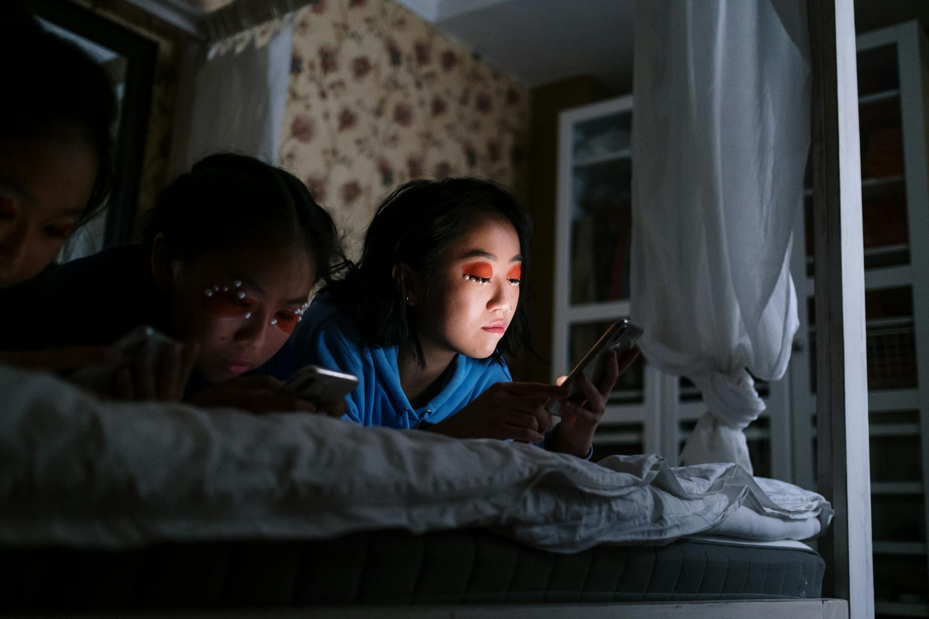 Three teenagers using smartphones in a bedroom at night, lit by their screens.