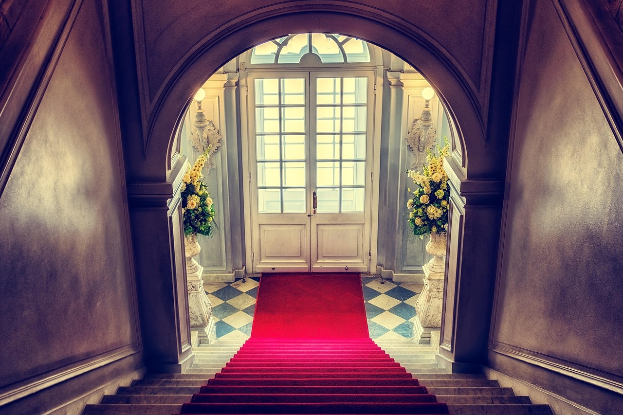 A grand staircase and red carpet inside a formal venue, evoking gala spectacle and social choreography.