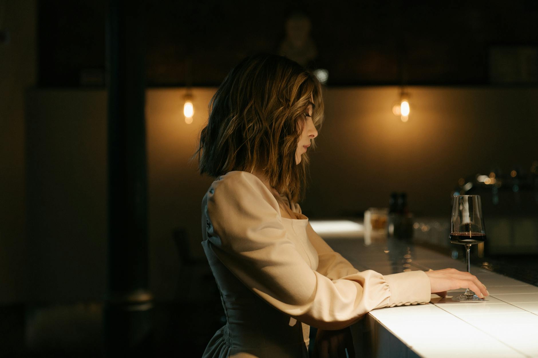 A woman sits alone at a warmly lit bar with a glass of wine