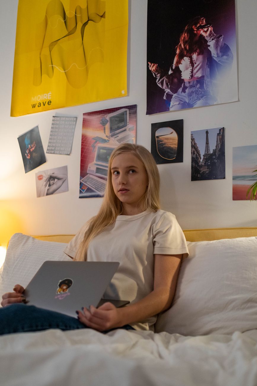 Teenage girl sitting on bed using laptop surrounded by various posters.