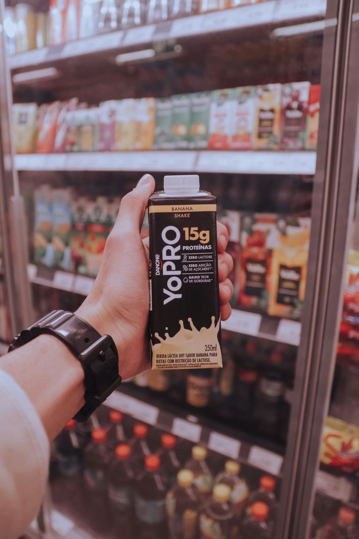 A hand holds a bottled protein shake in a supermarket aisle, the gym product now remade as an everyday convenience drink.